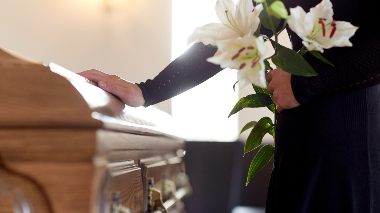 A man stands next to a casket with white flowers in hand.